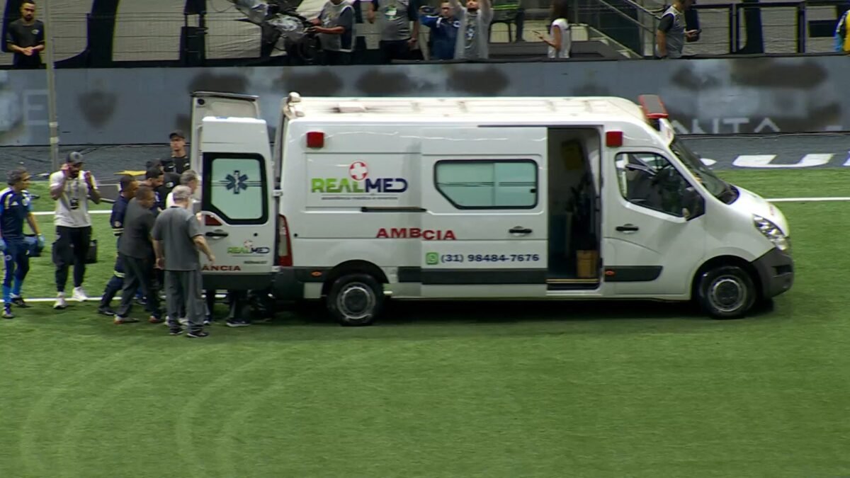 Ambulancia branca parada no gramado do estadio com portas abertas e equipe medica prestando atendimento