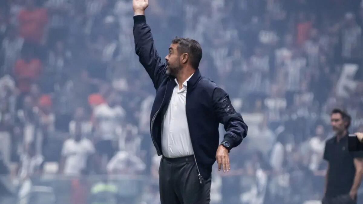 Treinador gesticula com o braço levantado à beira do gramado em estádio de futebol cheio