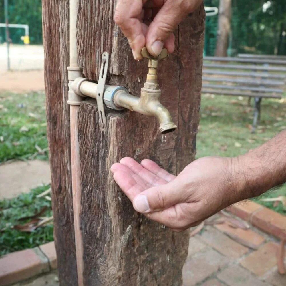 Pessoa tenta abrir uma torneira de metal instalada em um tronco de madeira com as mãos em concha embaixo