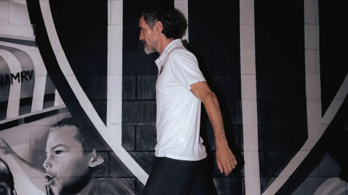 Homem de perfil vestindo camisa branca e calça preta caminha diante de parede com pintura em preto e branco