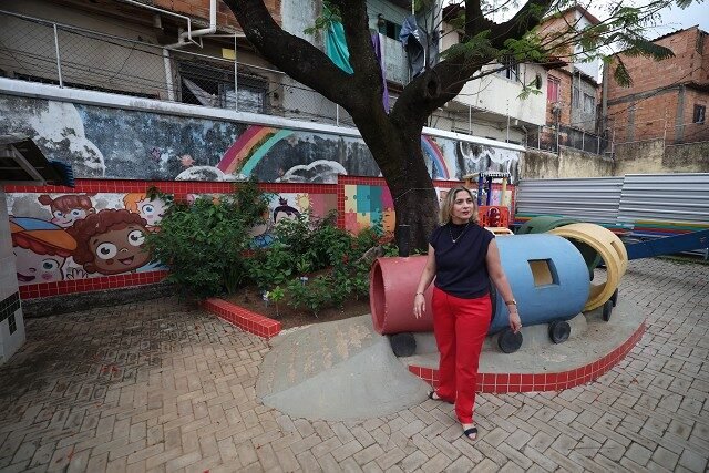 Mulher adulta de calça vermelha e blusa preta em pé no pátio de escola municipal em Belo Horizonte
