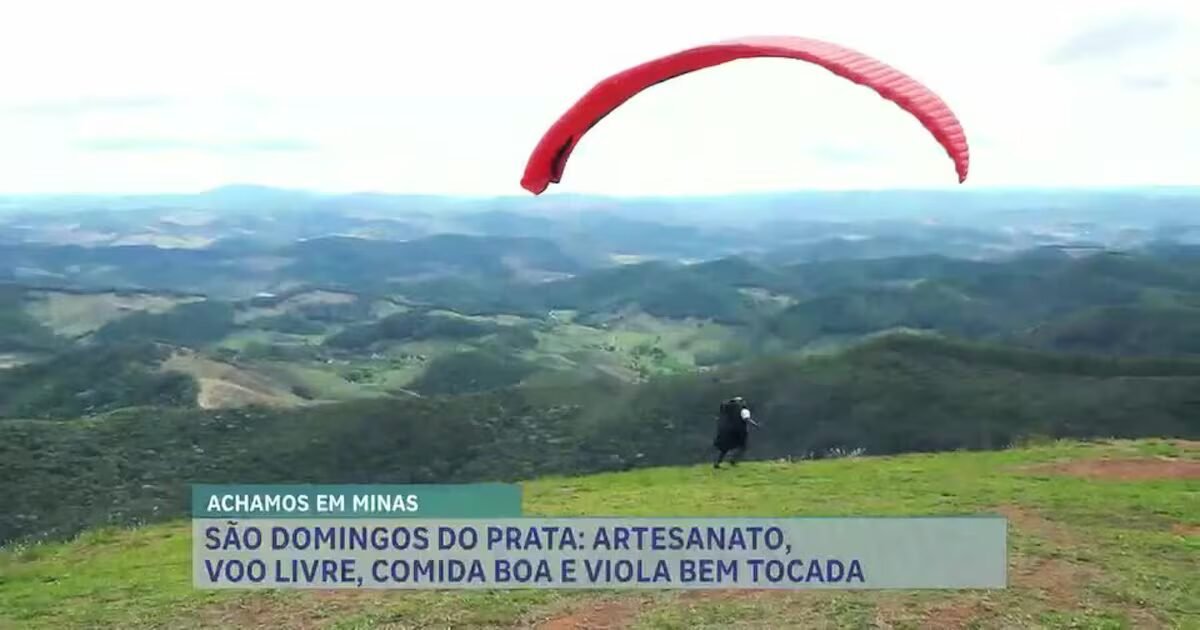 Pessoa se preparando para decolar de paraglider vermelho em encosta verde com vista para montanhas ao fundo