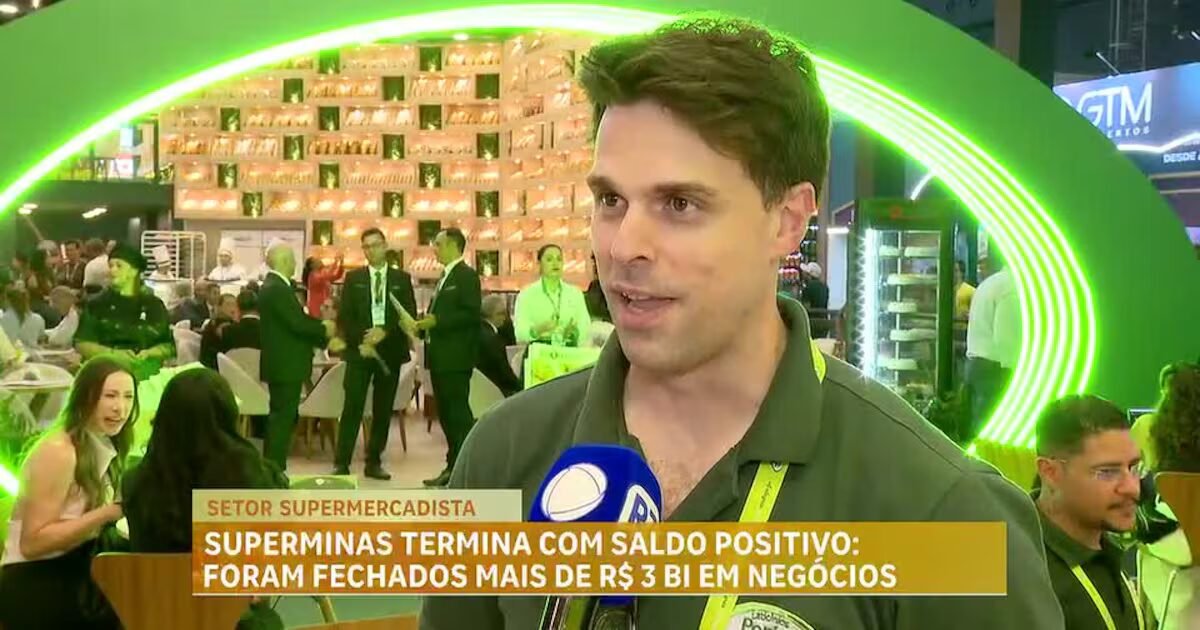 Homem de polo verde fala em microfone durante a Superminas com feira movimentada ao fundo